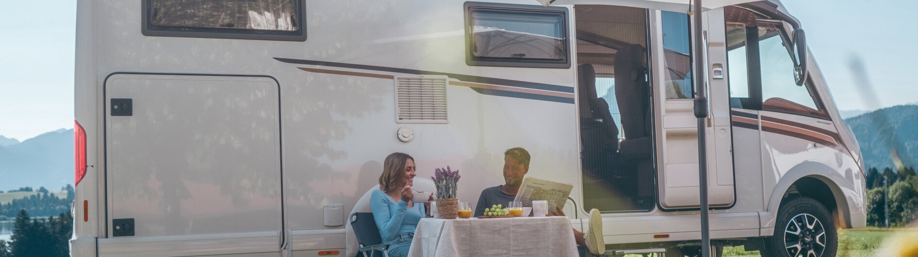A man and a woman sitting in front of their campervan under a sunshade, drinking coffee.